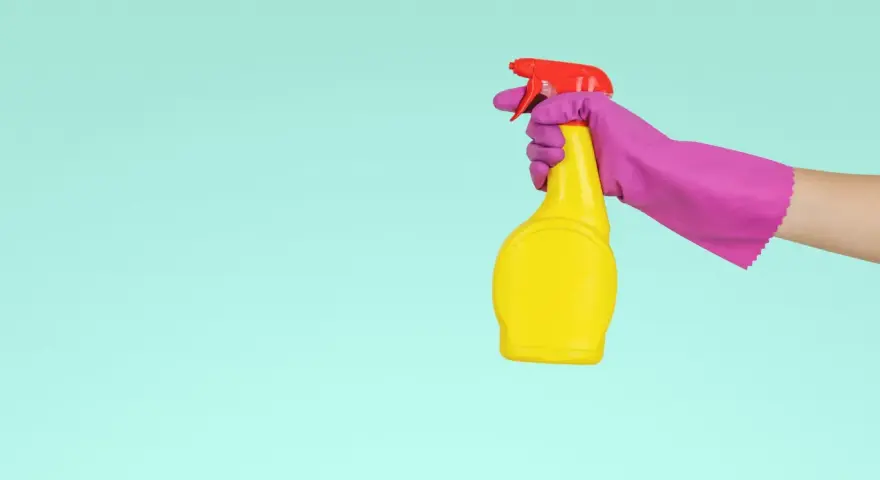 Main tenant, une main gantée de rose tient un flacon jaune prêt à nettoyer le sol en vinyle avec du vinaigre blanc.
