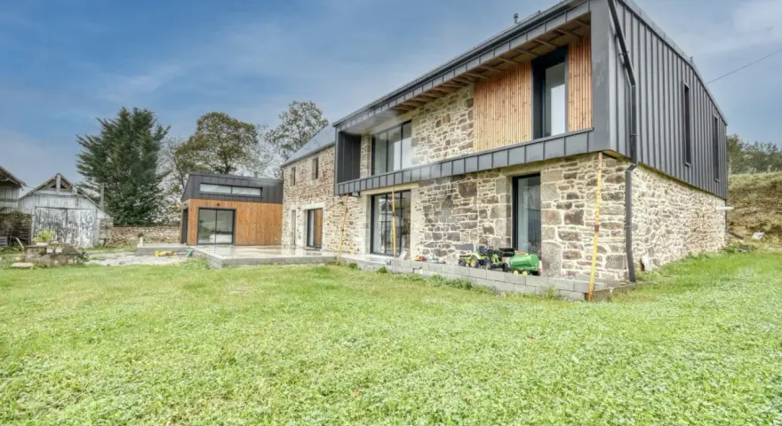 Une maison en pierre rénovée, mêlant tradition et modernité avec des touches de bois et de métal. Un jardin verdoyant entoure cette demeure.