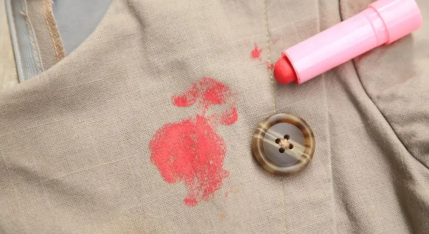 Rouge à lèvres sur polyester : une tache rouge vif sur un tissu beige, à côté d'un bouton marron et du tube de rouge à lèvres rose. Comment enlever cette tache ?