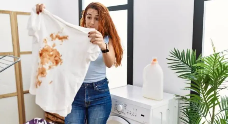 Femme surprise devant un t-shirt blanc taché de marron, prête à détacher le linge blanc. Machine à laver et lessive à proximité.