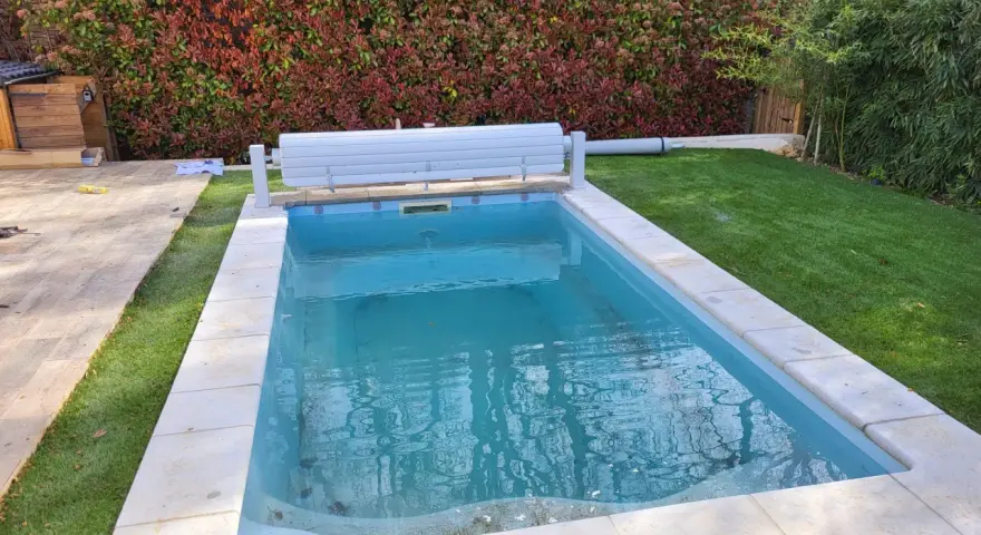 Piscine en béton avec des taches noires sur le fond, entourée de gazon et d'une haie.