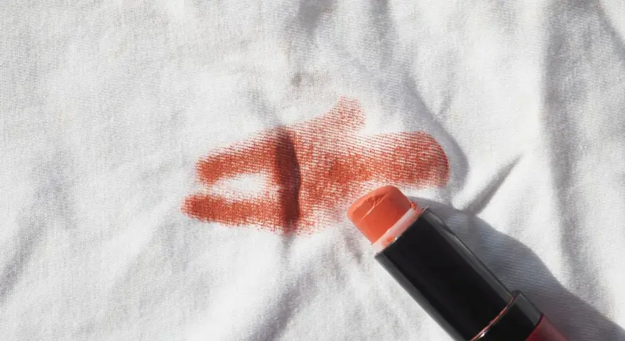 Un bâton de rouge à lèvres orange est posé sur un tissu blanc, laissant une tache rouge à lèvre.