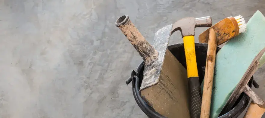 Outils pour nettoyer du ciment noirci : marteau, truelle, brosse et éponge sur fond de béton.