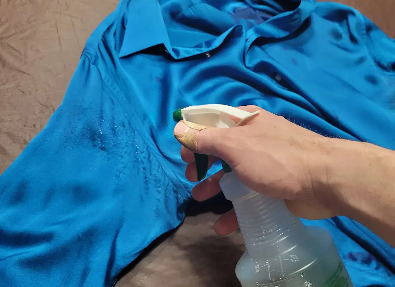 Une main avec un pansement sur le pouce vaporise un produit sur une chemise bleue, tentant d'enlever une tache de gras sur soie.