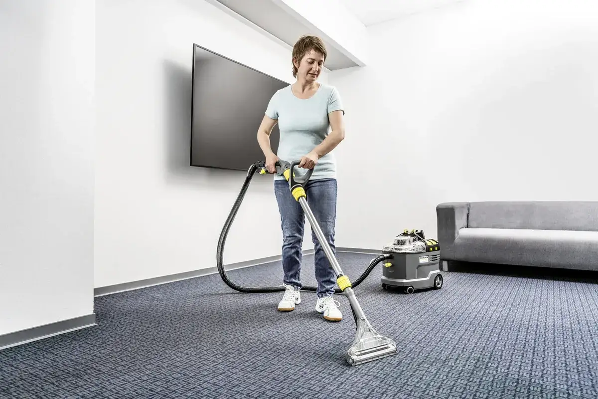 Une femme utilise un appareil pour le nettoyage moquette vapeur. Un canapé gris et un écran plat sont visibles.