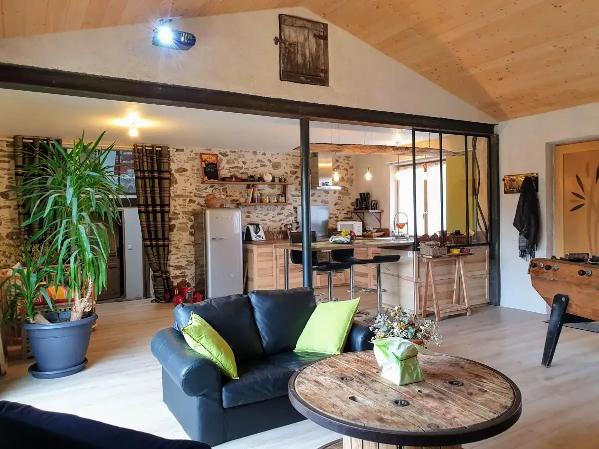 Salon cosy d'une maison en pierre avec canap&eacute; noir, table basse en bobine de c&acirc;ble, et cuisine ouverte.
