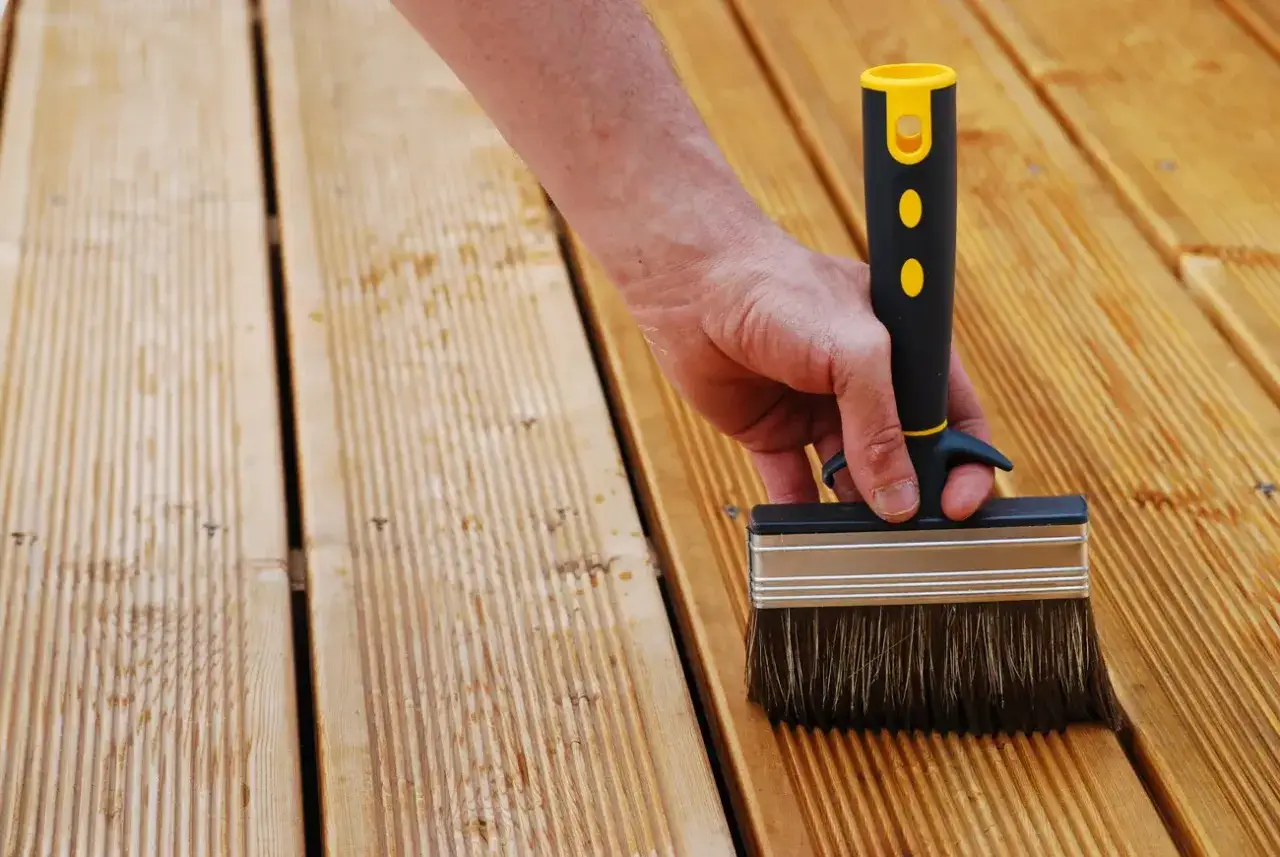Main tenant une large brosse pour enlever le saturateur bois sur une terrasse en bois.
