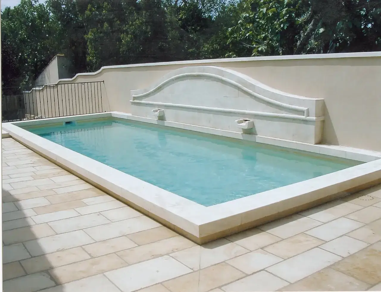 Une piscine rectangulaire aux marges en pierre, entour&eacute;e d'un dallage clair. Un mur orn&eacute; d'une fontaine en pierre domine le bassin.