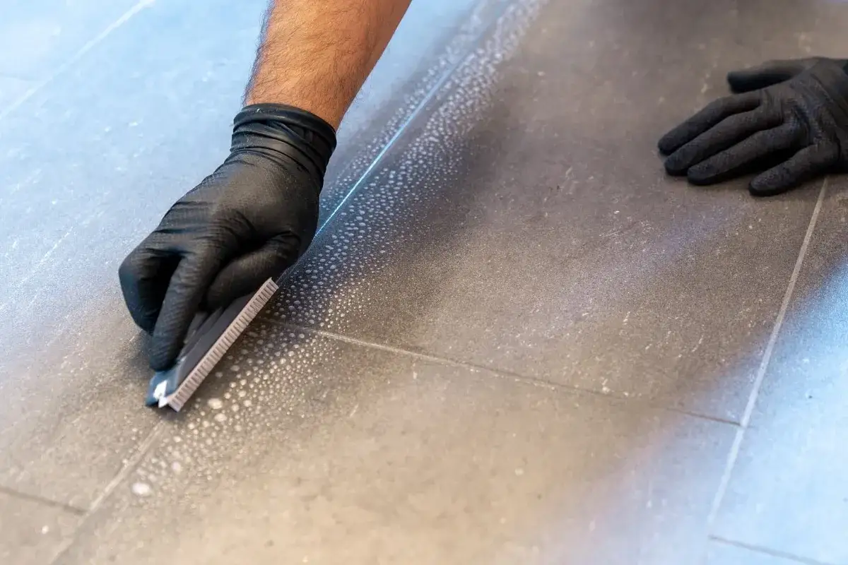 Main tenant avec des gants noirs, utilise un grattoir pour enlever du joint de carrelage sec.