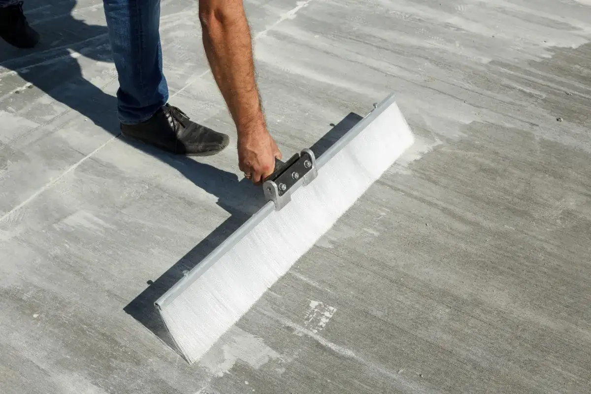 Un homme utilise une brosse large pour rénover une terrasse en dalle béton, préparant la surface pour un nouveau revêtement.