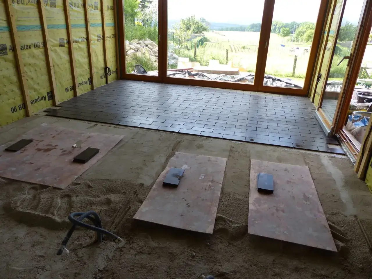 Pose de tomettes sur dalle béton. Le sol est en cours d'installation, avec des tomettes sombres posées sur une dalle de béton, et des plaques de protection sur le sable.