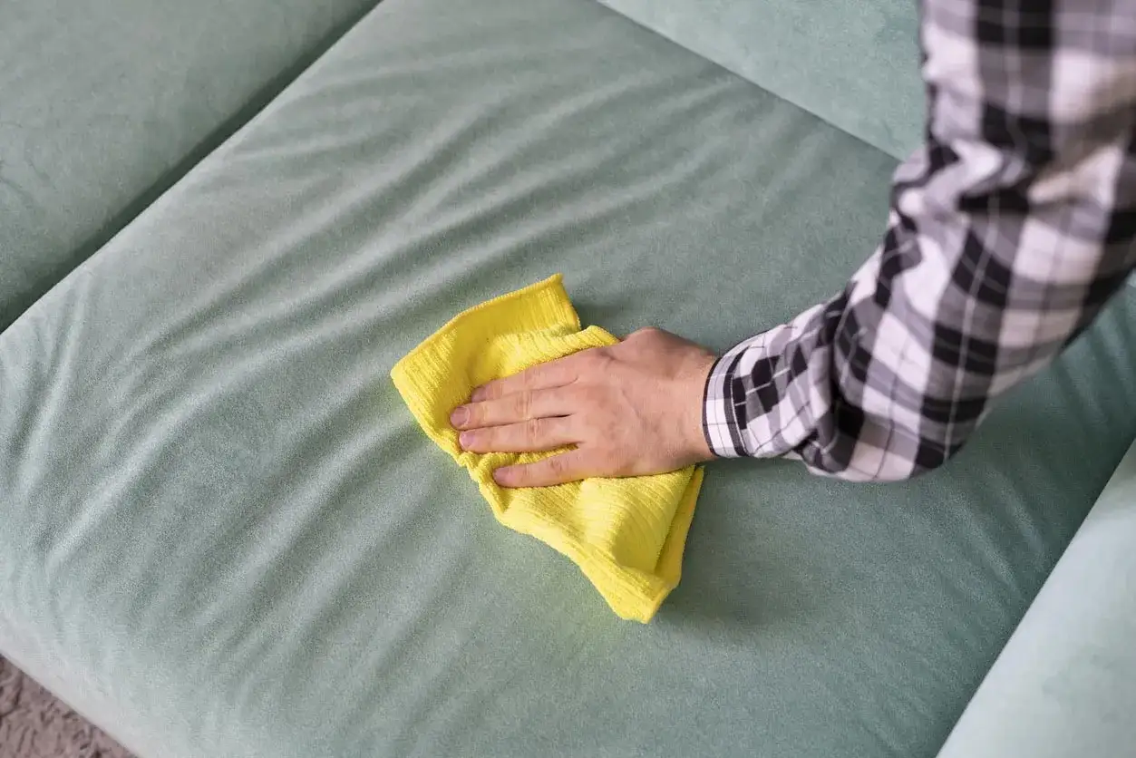 Main d'une personne nettoyant un canapé en tissu avec un chiffon jaune. Une astuce pour nettoyer canapé tissu vinaigre blanc bicarbonate.
