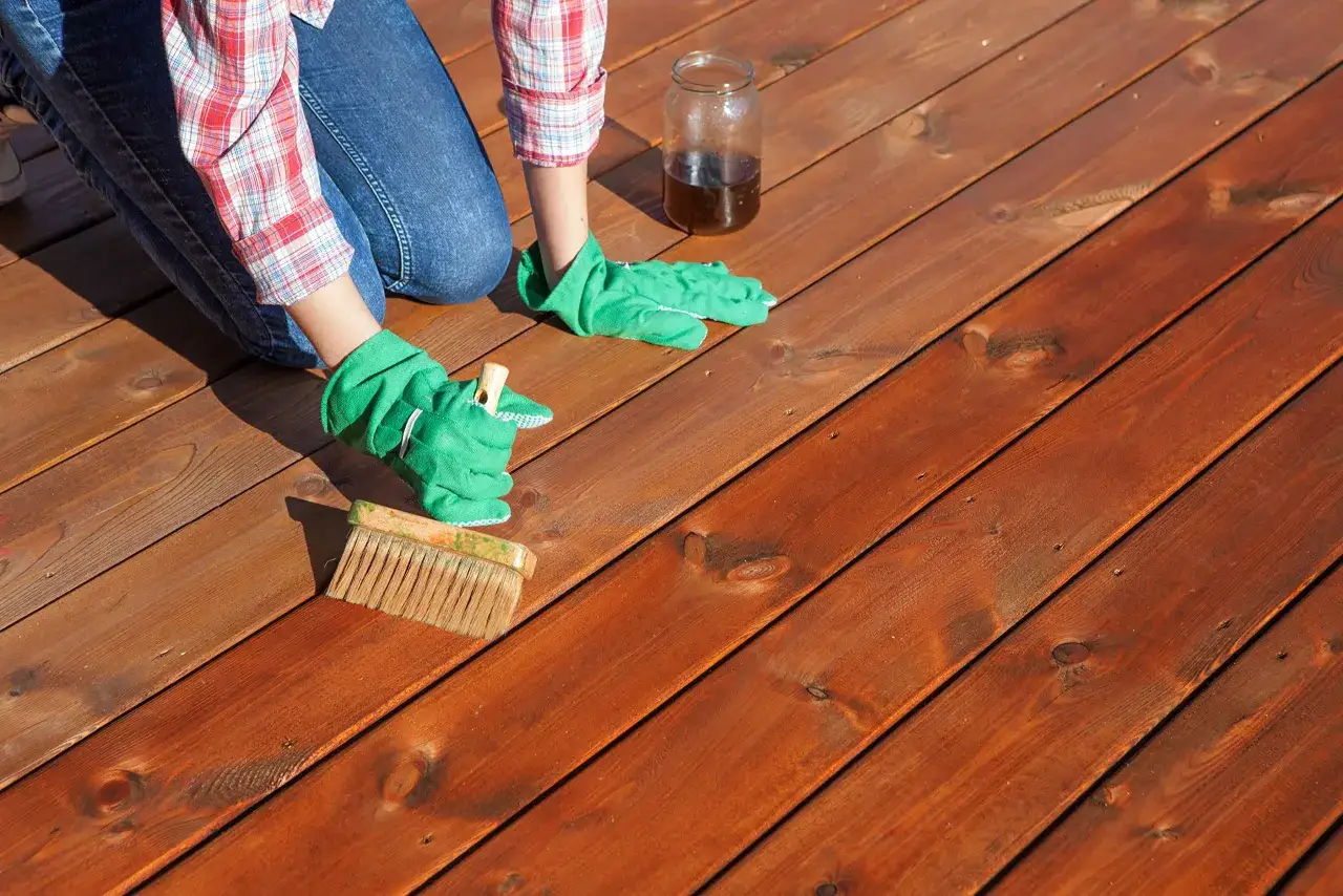 Application d'une huile pour fabriquer son huile de teck sur une terrasse en bois. Une personne en gants verts utilise un pinceau.