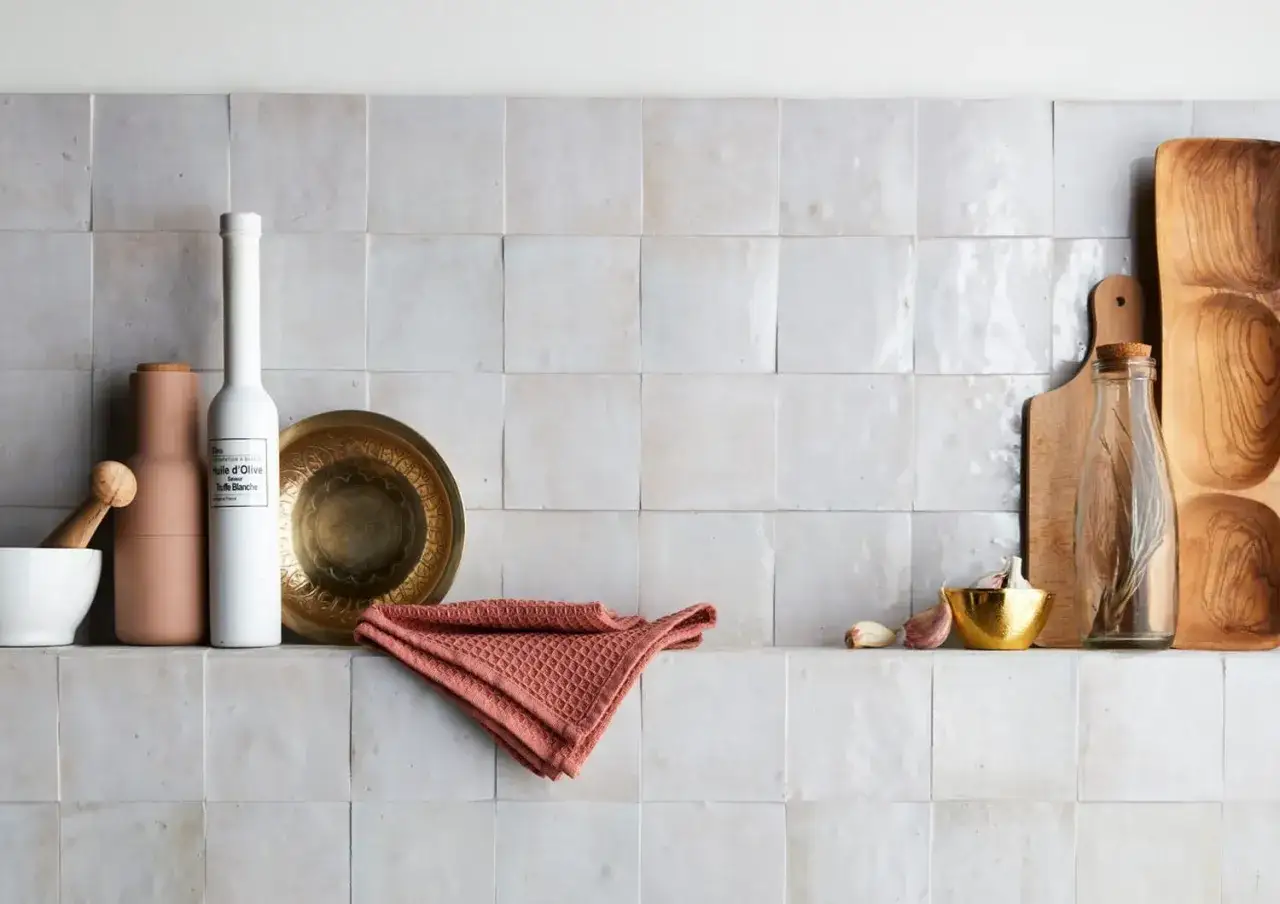 Sur un mur en carrelage poreux, des ustensiles de cuisine : mortier, moulin, bouteille d'huile d'olive, plat en laiton, serviette corail, ail, et planche &agrave; d&eacute;couper en bois.