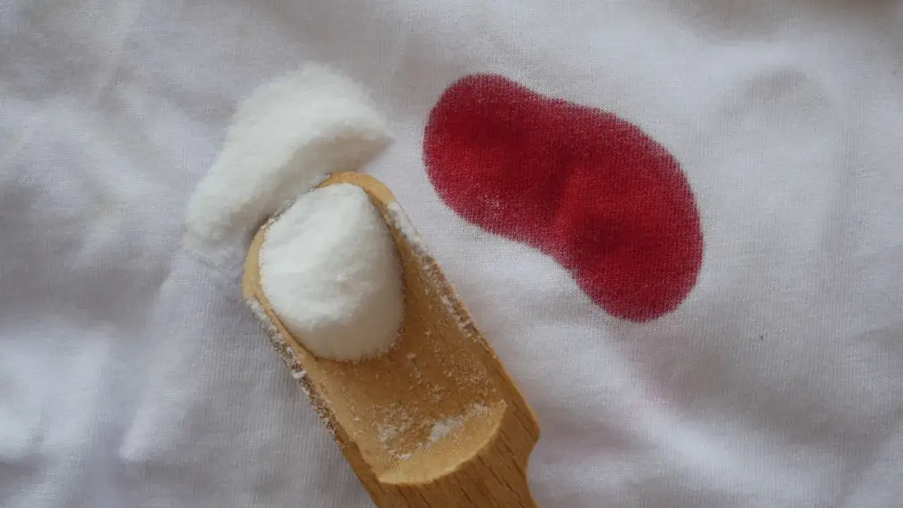 Une tache rouge sur du coton blanc, &agrave; c&ocirc;t&eacute; d'une cuill&egrave;re en bois remplie de poudre blanche, sugg&eacute;rant comment enlever des taches de sang sur du coton.