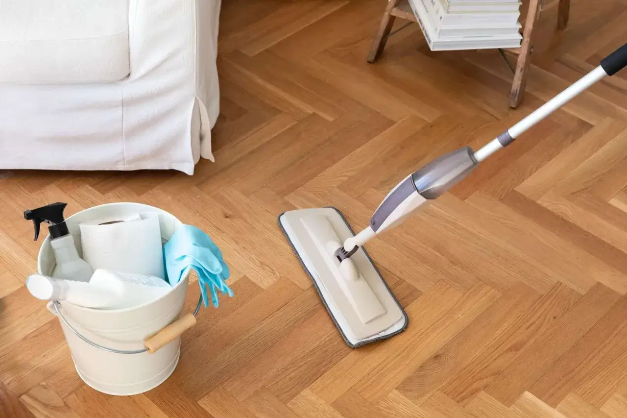 Nettoyer sol vinyle vinaigre blanc : un seau de produits ménagers et une serpillière à pulvérisation sur un parquet en chevrons.