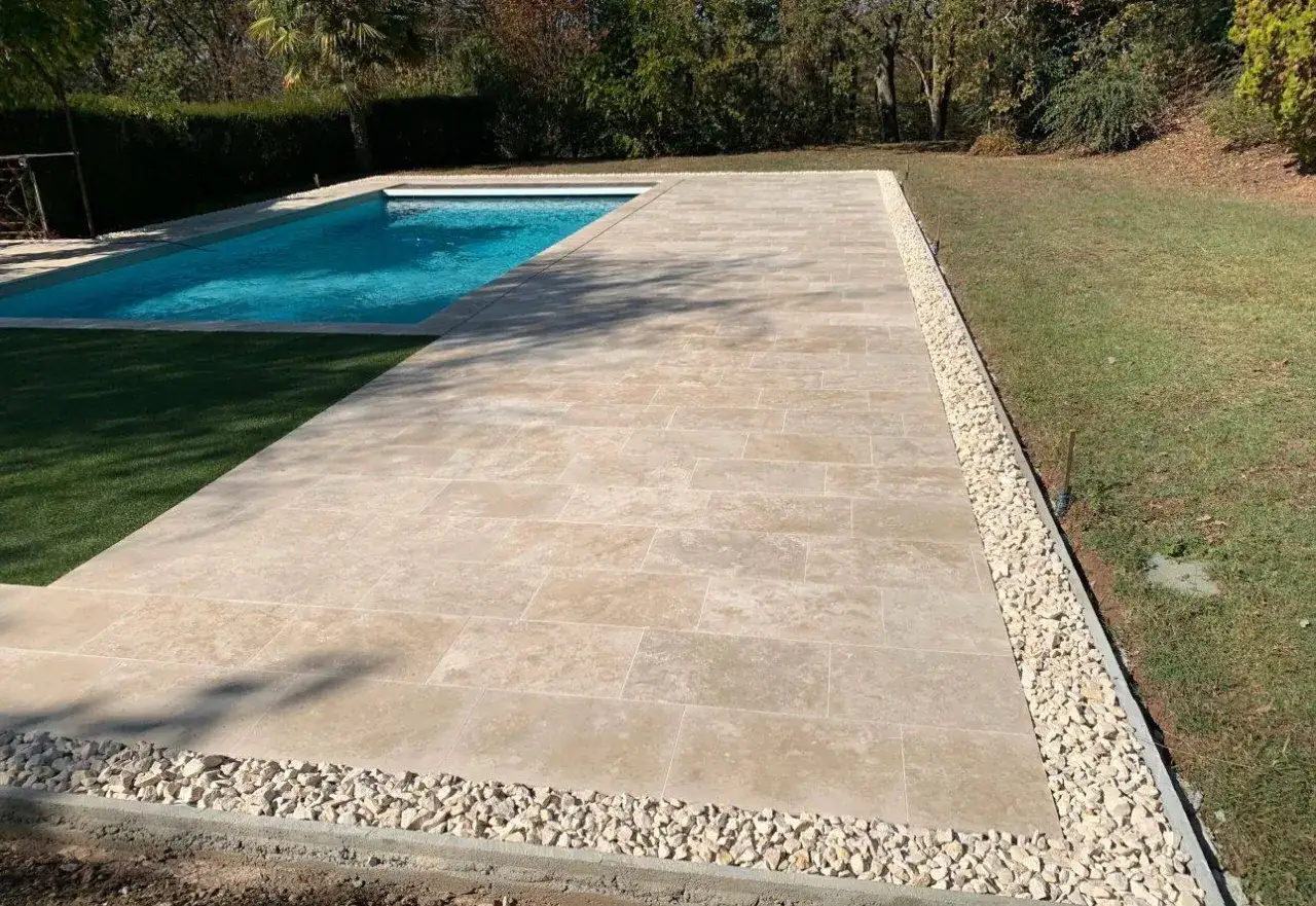 Une belle terrasse en travertin autour d'une piscine bleue, bord&eacute;e de gravier blanc et de pelouse verte.