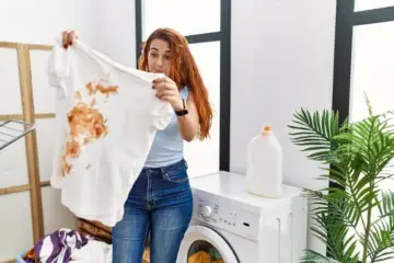 Femme surprise devant un t-shirt blanc taché de marron, prête à détacher le linge blanc. Machine à laver et lessive à proximité.